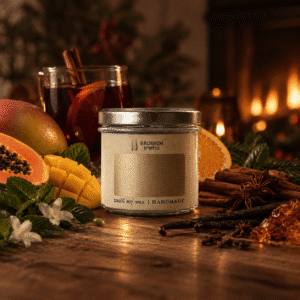 Натуральная соевая аромасвеча "Глинтвейн" (Mistletoe & Wine) от Broshim Candles в прозрачной стеклянной банке с зажженным деревянным фитилем. Свеча стоит на рустикальном деревянном столе в уютной зимней обстановке, окруженная палочками корицы, бадьяном, сушеными апельсинами, гвоздикой и веточками омелы. На фоне — размытые огни рождественской елки и теплый камин, создающие атмосферу праздника и уюта.